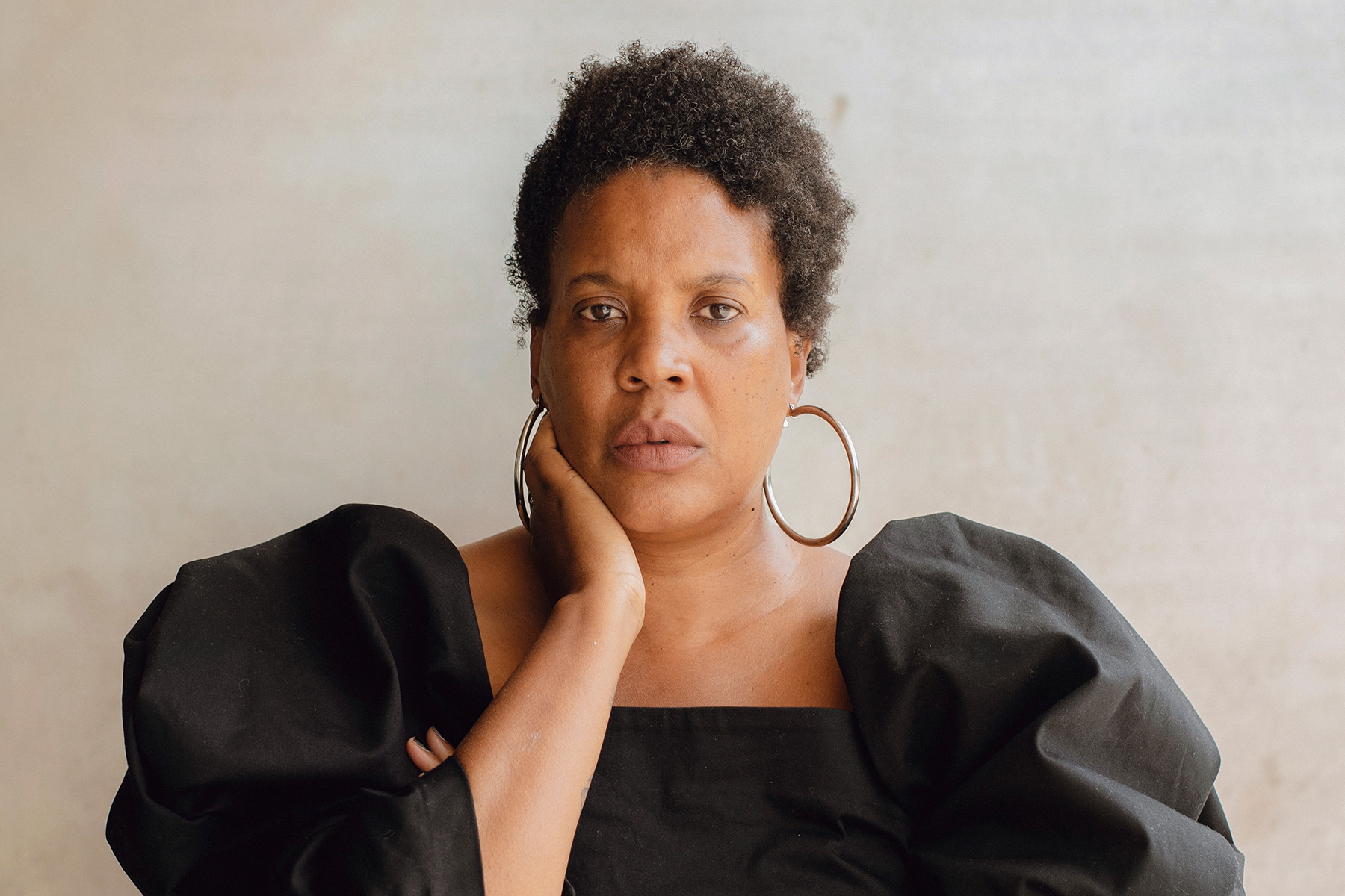 Image of a woman looking pensively into the camera, wearing a black dress and large hoop earrings, with one hand tucked behind her right ear.; Read More in the New York Times