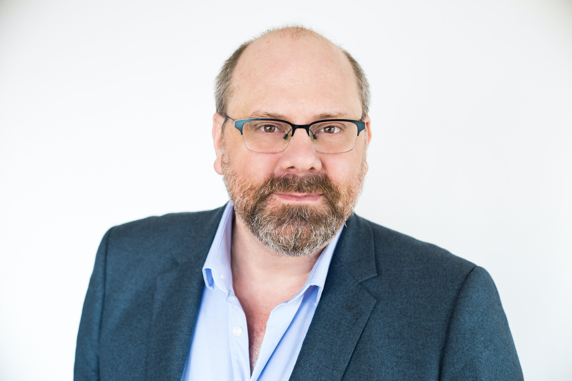 Image of a man looking thoughtfully into the camera, wearing a blue blazer, a blue button-down shirt, and black-framed glasses.; Read More in the New York Times