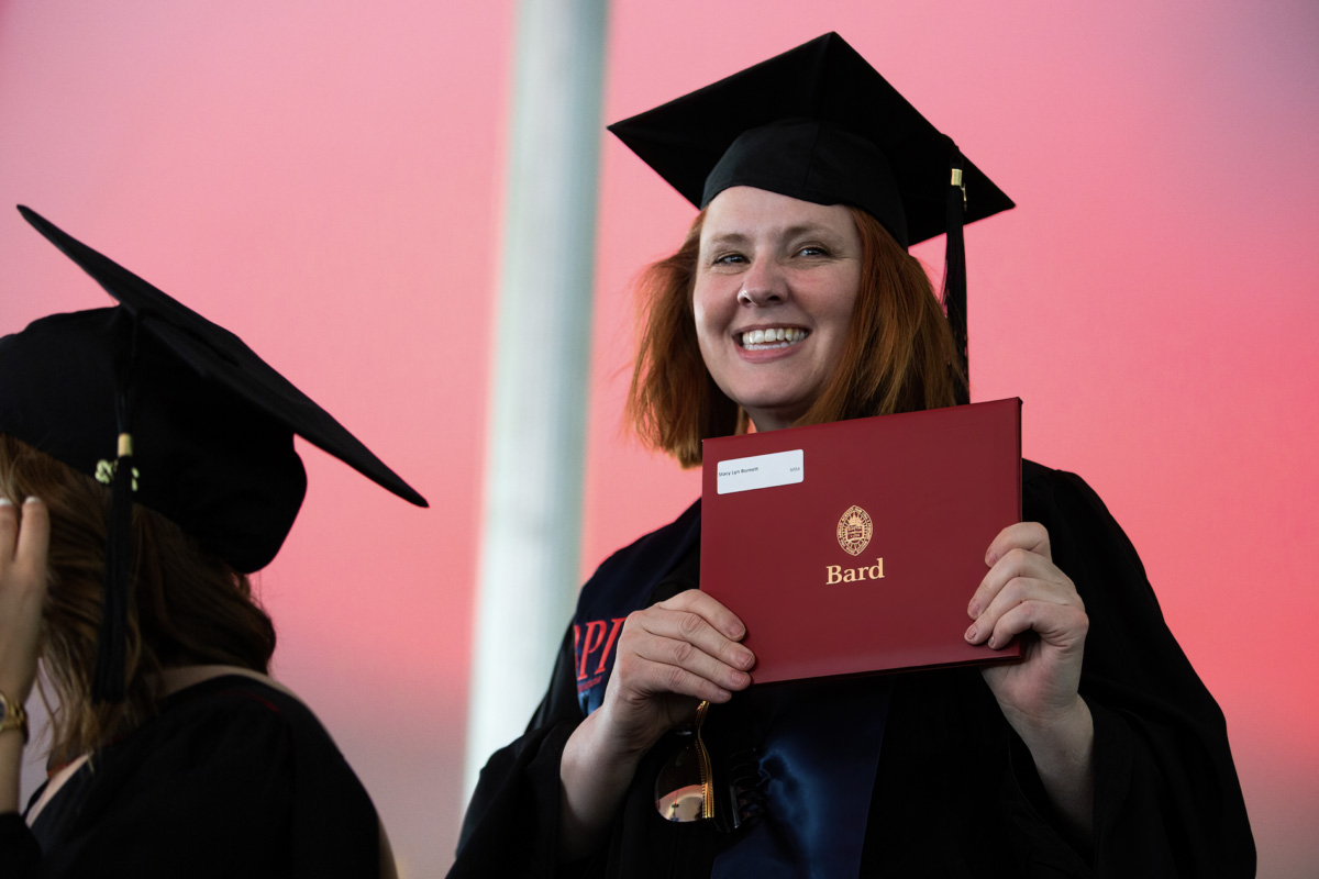 Smiling woman with red hair in cap and gown, holding up a red diploma for the camera.; Read More in Diverse Education