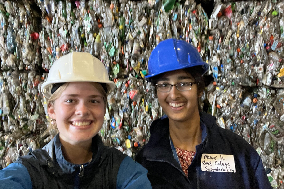 Two students wearing hard hats smile at the camera with a wall of recycled plastic materials behind them.; Learn More