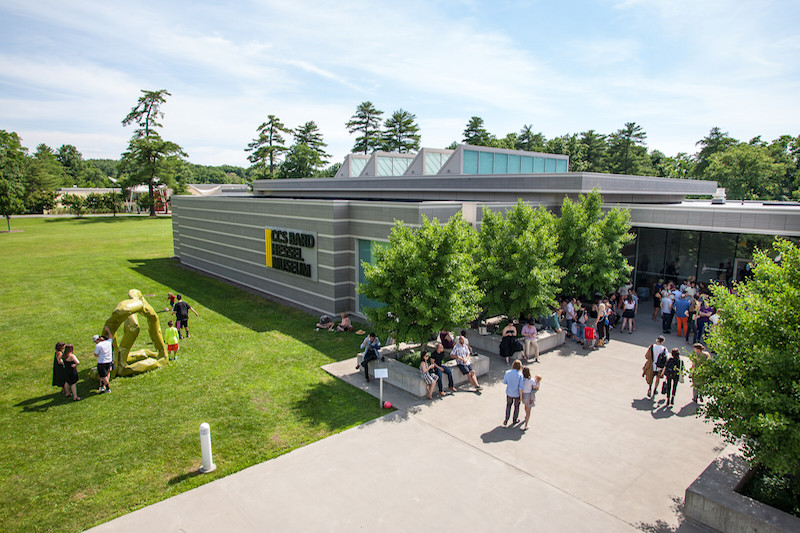 Exterior shot of the Center for Curatorial Studies, Bard College: a large, metal building surrounded by green grass and trees. People walk around on a concrete patio near the entrance.

