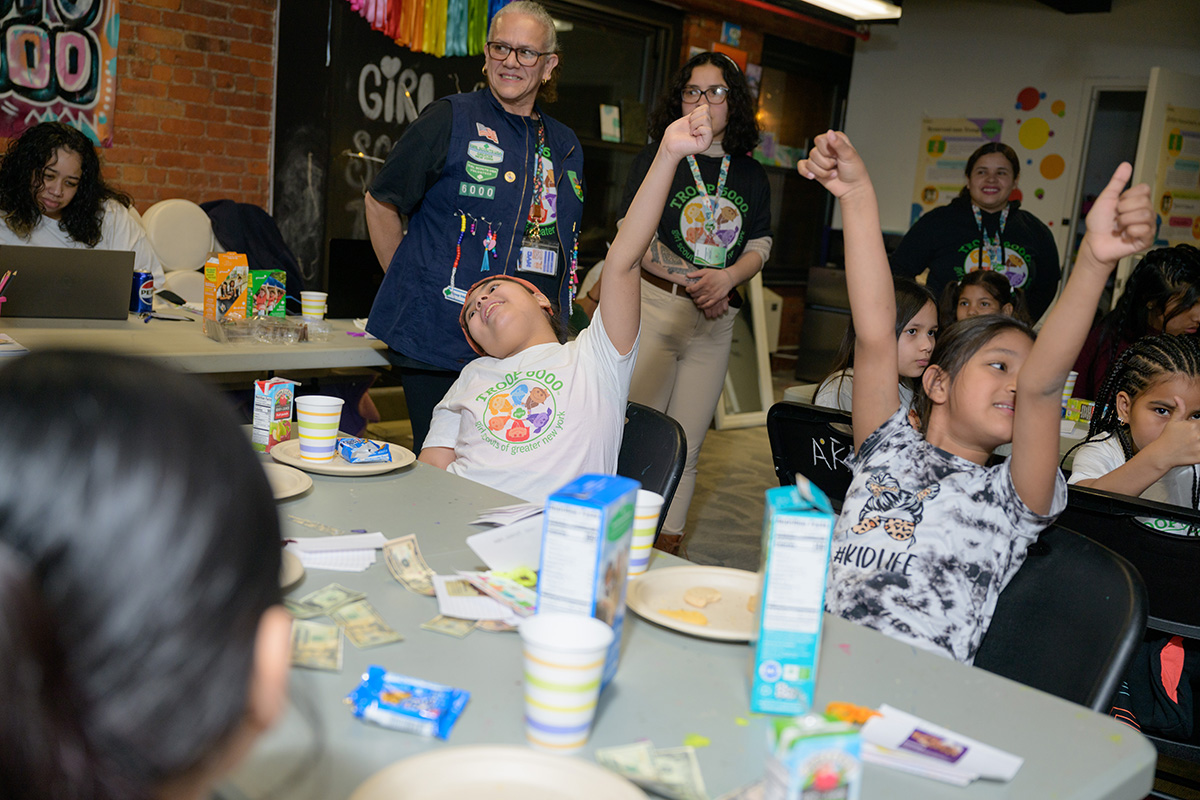 A group of young girls sitting at a table wearing Troop 6000 t-shirts raise their arms in the air and give the thumbs up.; Read More on NPR