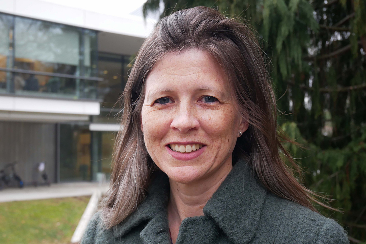A woman in a gray jacket with shoulder length brown hair smiles at the camera. Behind her is a silver academic building and green trees.