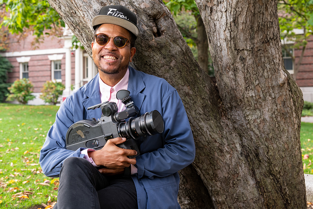 A man in a pink shirt, blue jacket, and sunglasses leans against a tree holding a camera meant for producing films.; Read More in&nbsp;ArtReview