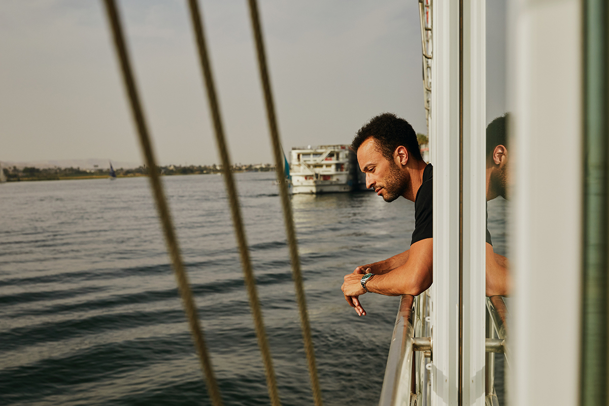 A man in a black t-shirt leans over the railing of a boat, the water in front of him as he looks down.