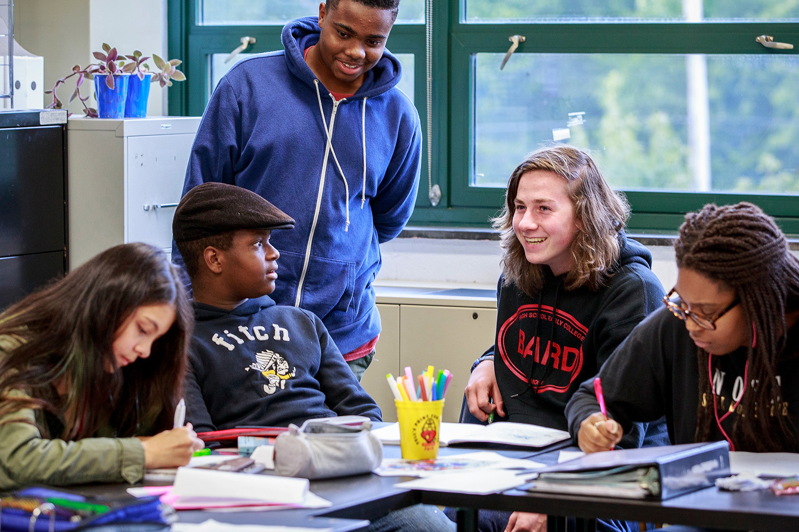 Five students sitting and standing around a table. Two students are writing and the rest talk.