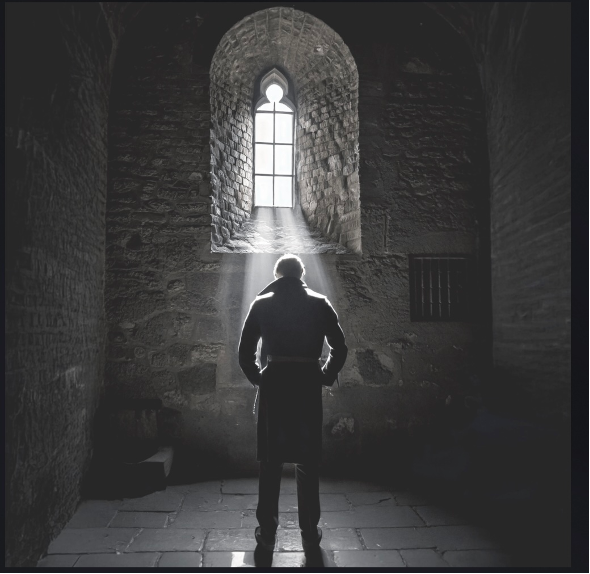 Man standing with back towards viewer and facing rays of light coming through window in old stone room. 