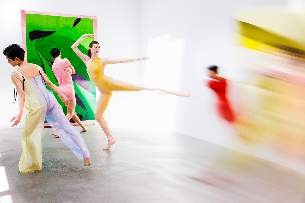 Four dancers in mid-pose dressed in colorful costumes against white walls with a single painting.