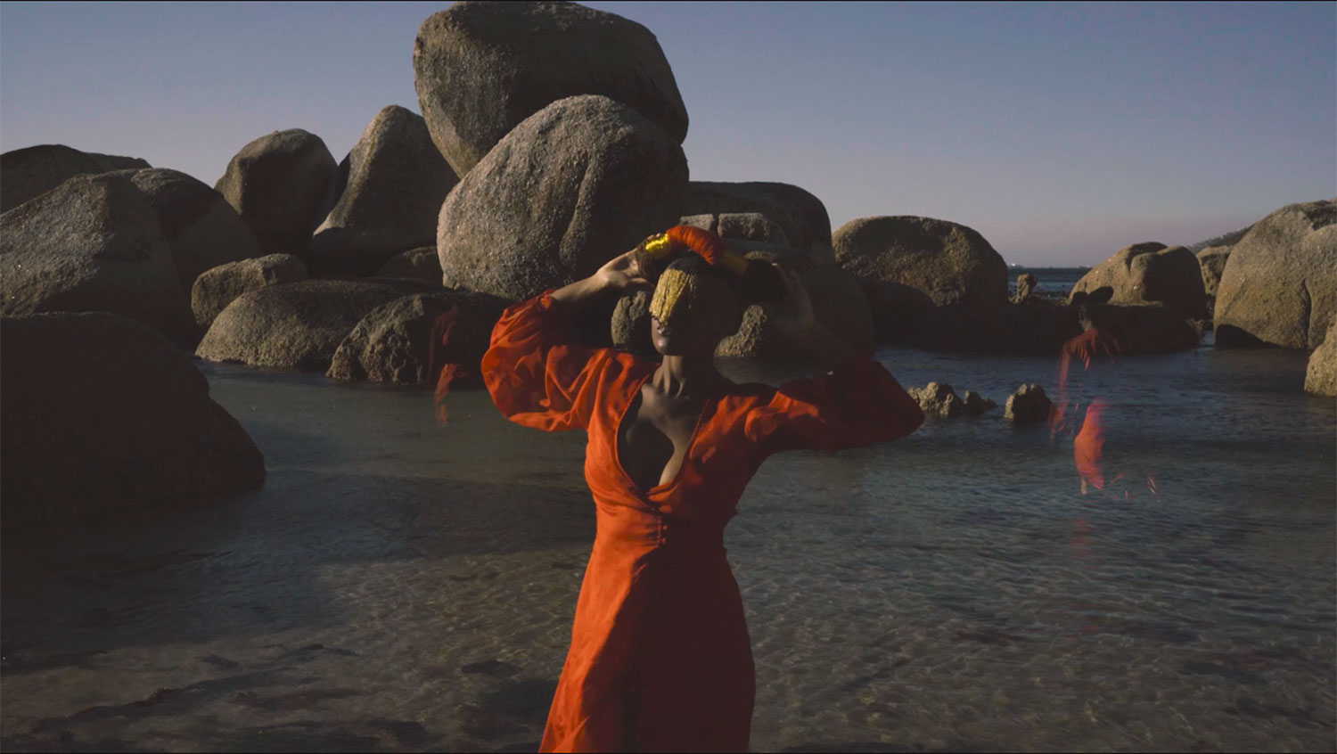 A woman in a red dress and golden veil appears on a coastal beach