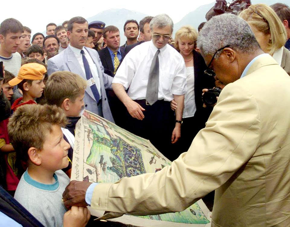 A young boy (Petrit Halilaj) shows his artwork to Kofi Annan, surrounded by a group of adults.