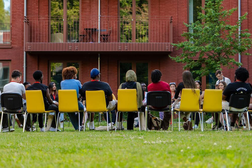 Students sitting in large circle of yellow chairs outside brick building.