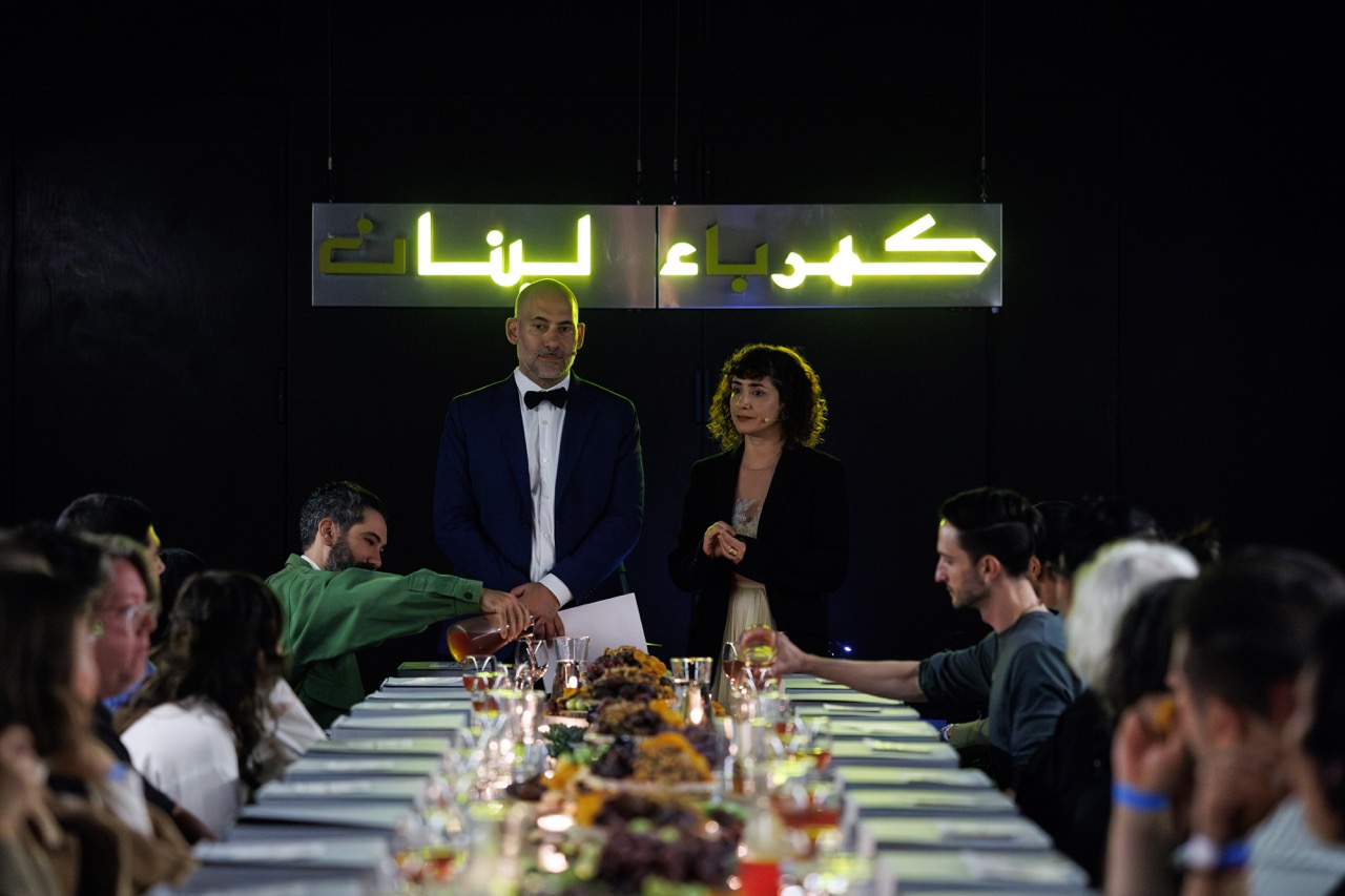 Ziad Abu-Rish and Tania El Khoury standing in front of an audience during The Search for Power performance. Text in arabic partially glows in neon yellow behind them.
