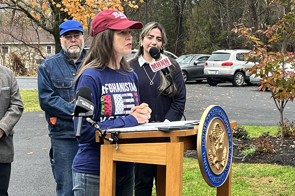 Woman wearing baseball cap speaking outside at podium.
