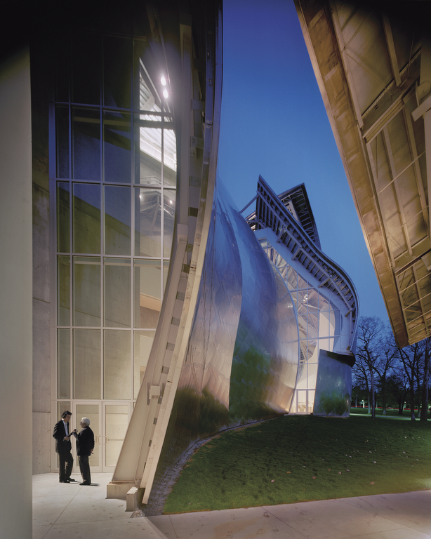 Two men speaking under the curved eaves of the stainless steel roof of a concert hall at twilight.