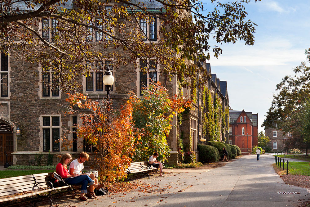 Students sitting on benches among college campus buildings of goth architecture surrounded by fall foliage.&nbsp;
