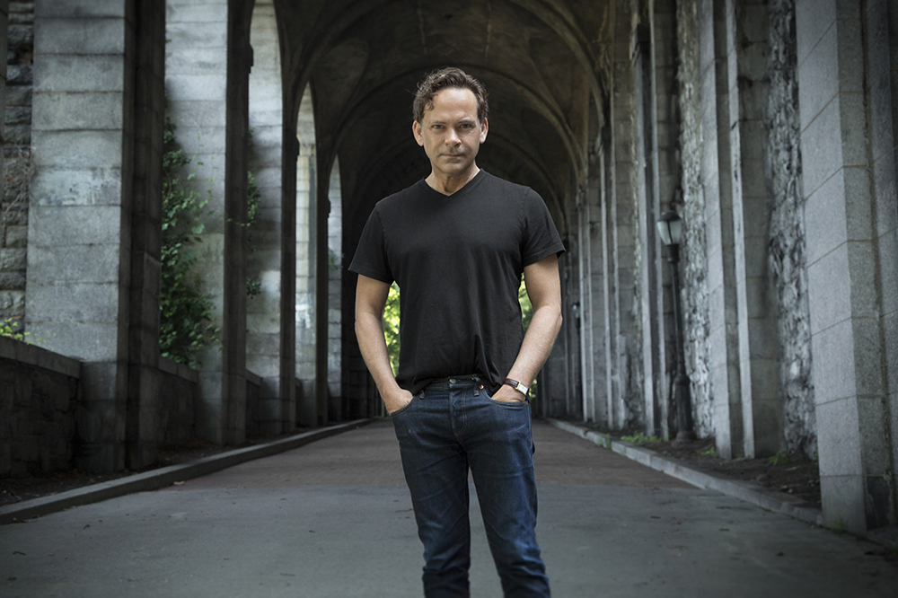 a man in a black t-shirt stands in front of a hallway of gothic stone arches