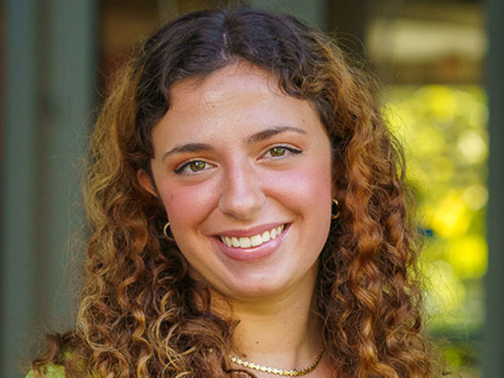 a woman smiles at the viewer against a backdrop of greenery