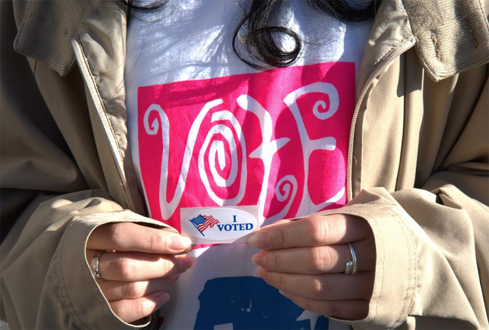 A person holding an "I Voted" sticker in front of their shirt which says "Vote."